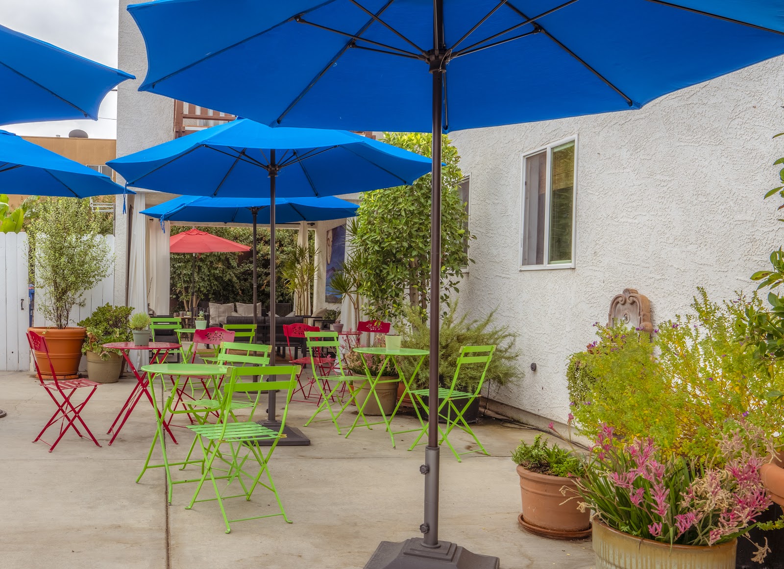 Exterior courtyard with blue umbrellas, colorful folding chairs, flowering plants, and white building walls