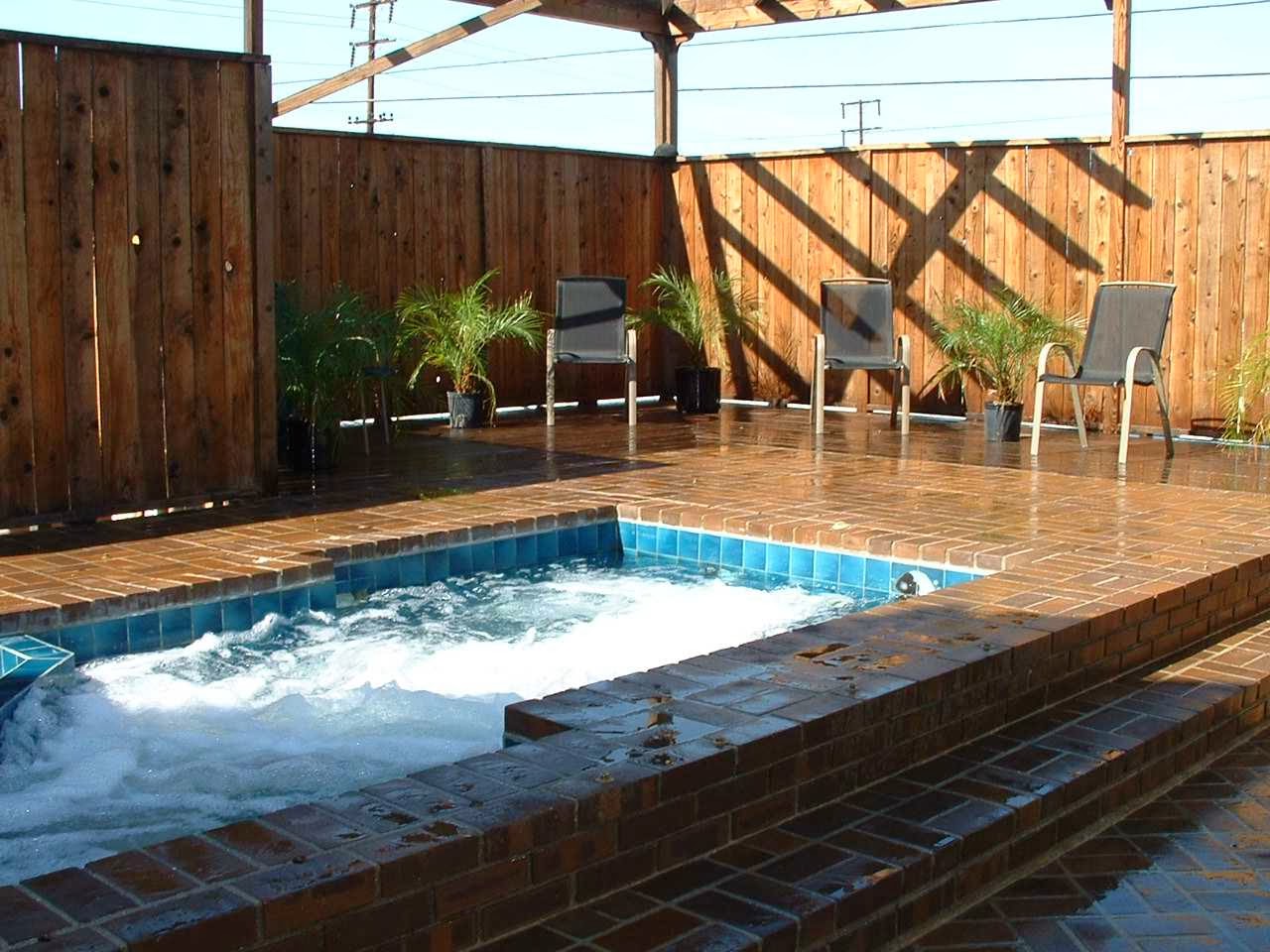 Outdoor patio with in-ground hot tub, wooden fence, pergola, lounge chairs, and brick paved deck area