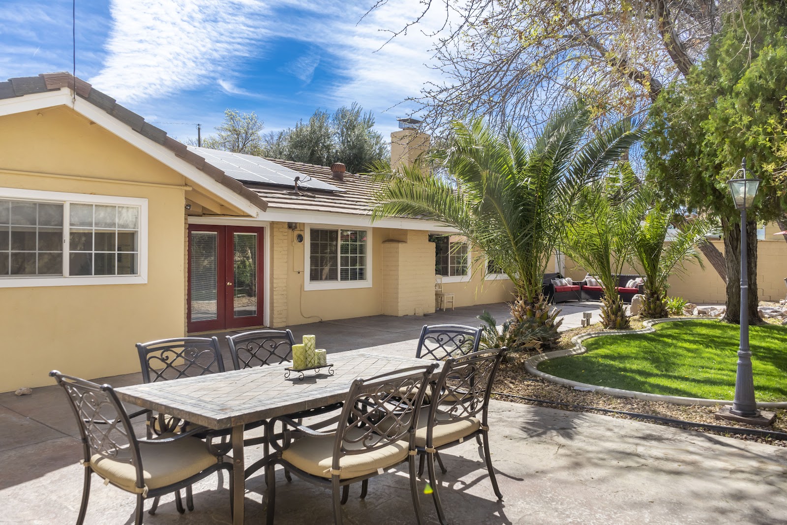 Outdoor patio with metal dining table and chairs, yellow building with red entry door, palm trees, and manicured lawn.