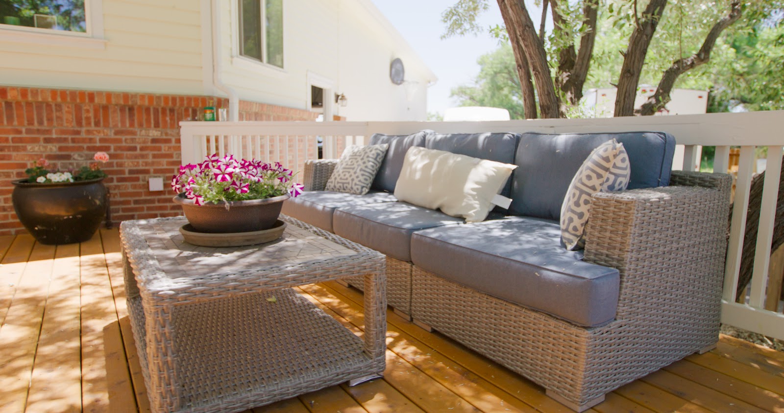 Outdoor brick patio with rattan woven seating, sectional sofa with cushions, glass-top table, and flowering plants.