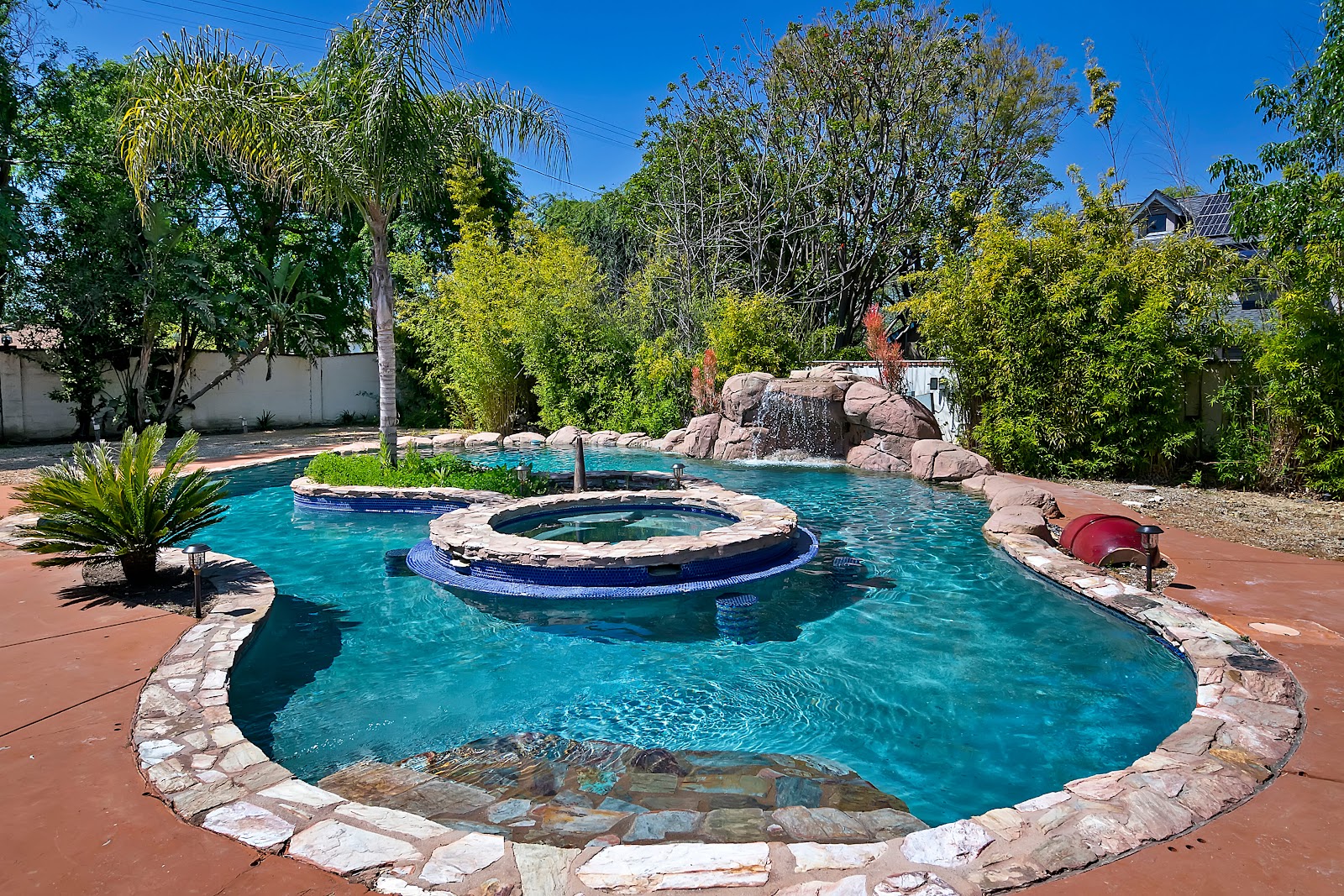 Exterior resort-style swimming pool with spa feature, stone decking, palm trees, lounge seating, and lush landscaping