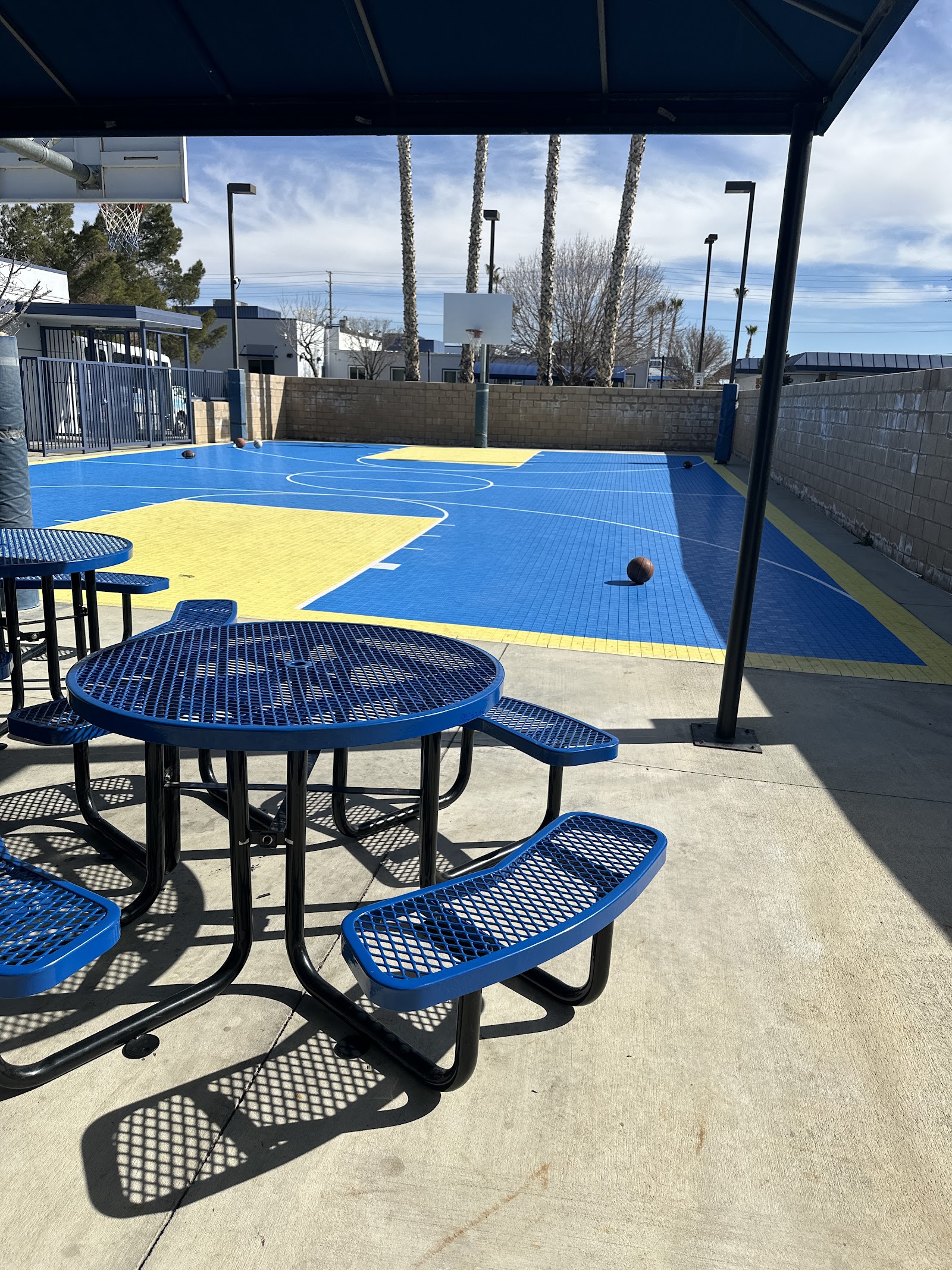 Exterior covered recreation court with blue and yellow basketball court markings, picnic tables with metal seating, palm trees visible beyond