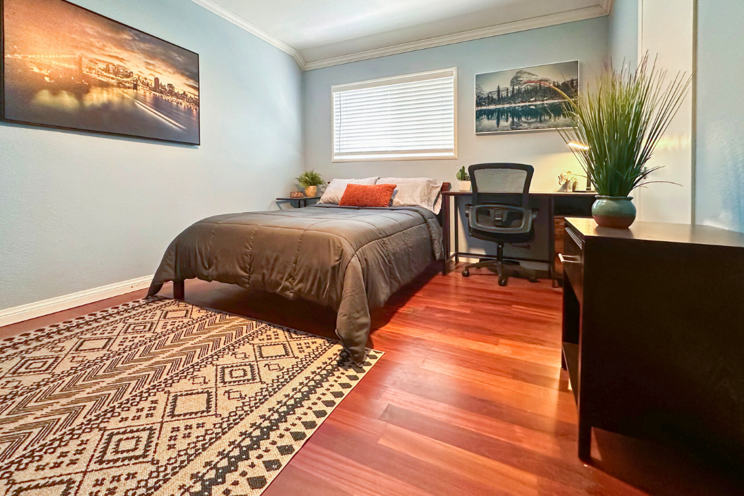 Interior patient bedroom with queen bed, desk, window with natural light, and hardwood flooring throughout.