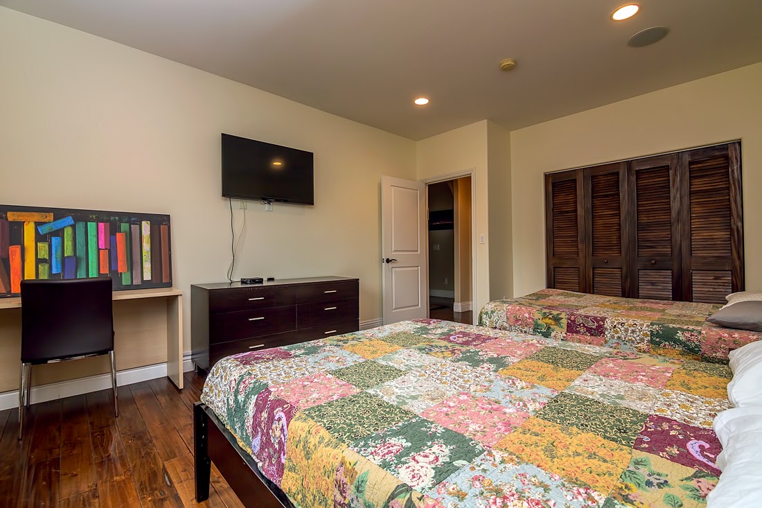 Interior patient bedroom featuring twin beds with patterned quilts, flat-screen TV, dark wood furniture, and modern lighting