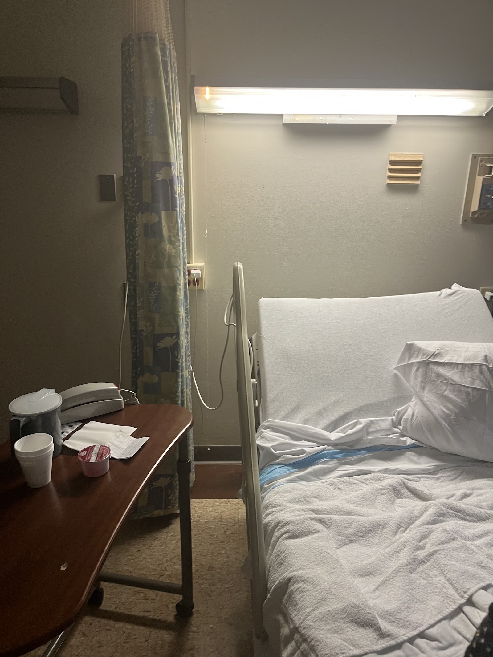 Interior hospital room with patient bed, white linens, bedside table, medical curtain, and wall-mounted hospital equipment