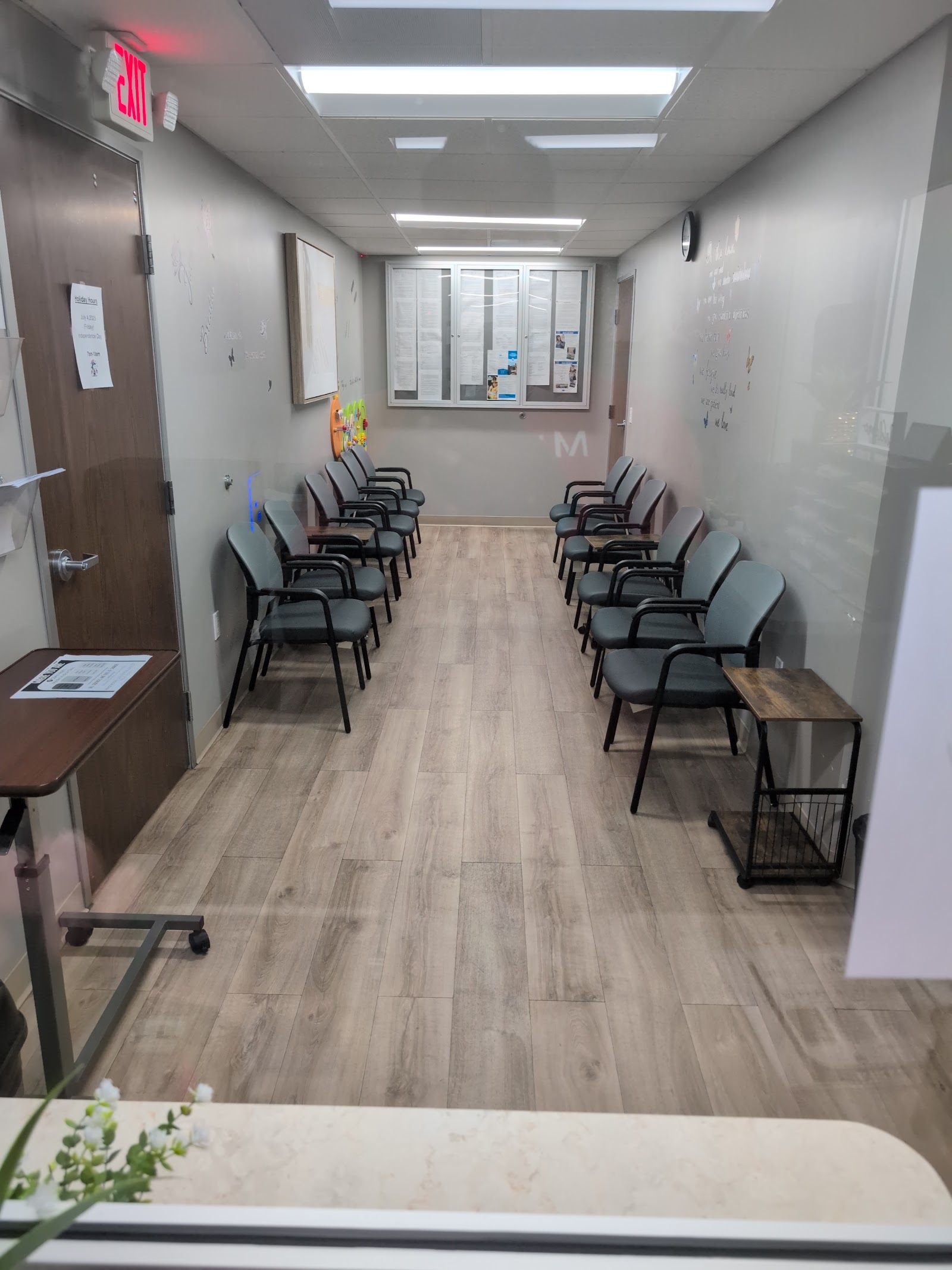 Interior waiting area with multiple black padded chairs arranged in two rows, whiteboard, window, and exit sign