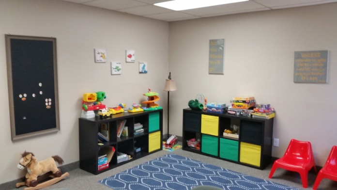 Interior playroom featuring black storage cubbies with toys, blue patterned rug, red chairs, and wall decorations.
