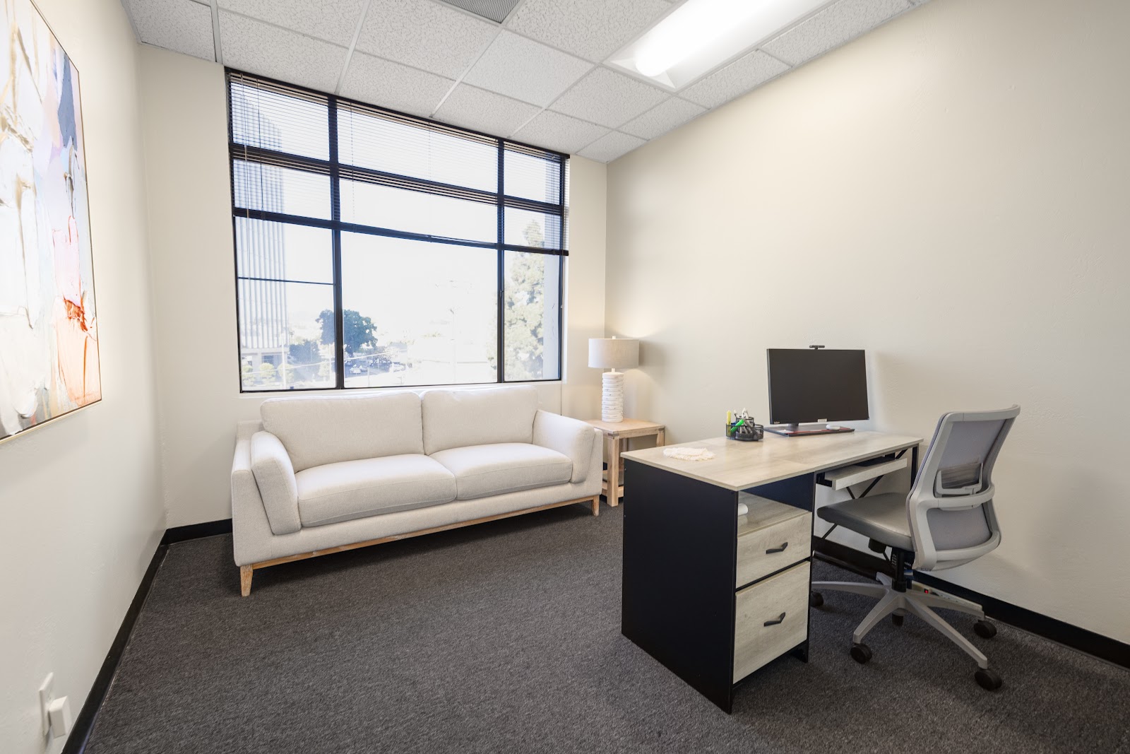 Interior office space with sofa, desk, computer monitor, office chair, and large gridded windows with natural lighting