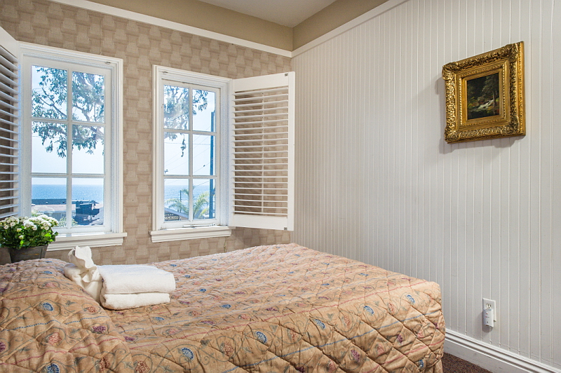 Interior bedroom with queen bed, ocean view windows with shutters, beige patterned wallpaper, white vertical paneling, and framed artwork