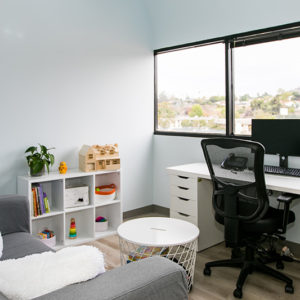 Interior therapy room with large window, storage shelving with white organizational bins, office chair, and small round ottoman seating