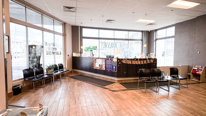 Interior reception area with light wood flooring, large windows, desk counter, seating chairs, and facility signage
