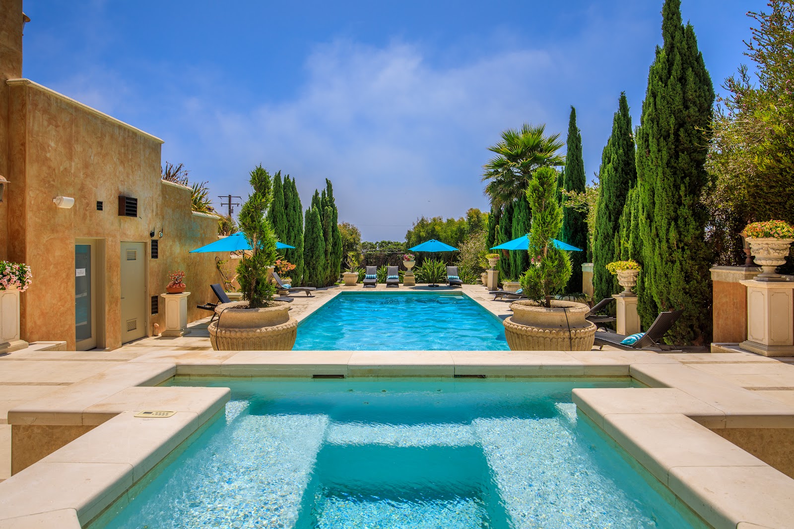 Exterior view of resort pool with lounge chairs, cypress trees, blue umbrellas, and Tuscan-style architecture