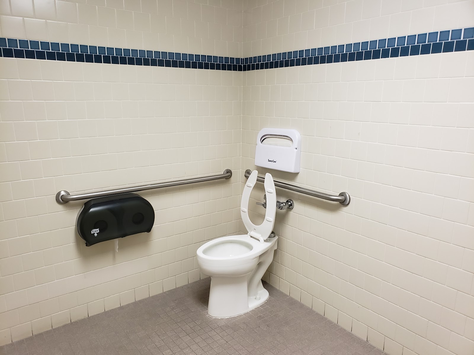 Interior restroom with white tile walls, accessible grab bars, toilet, sink, and paper towel dispenser