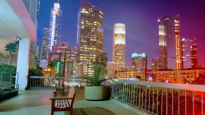 Exterior rooftop terrace at night featuring seating areas, planters, and metal railings with panoramic Los Angeles downtown skyline.
