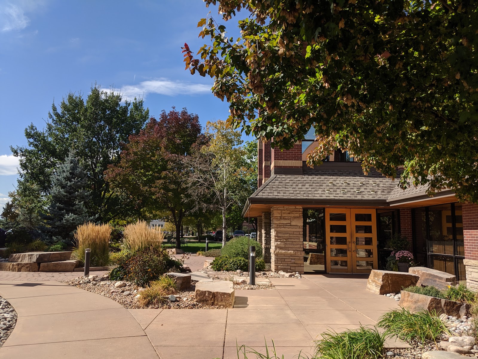 Exterior view of building entrance with stone seatwall, mature trees, decorative landscaping, and wooden glass doors