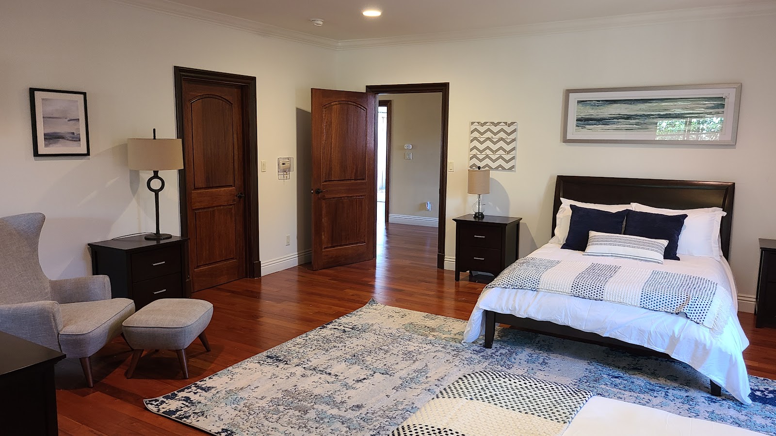 Interior bedroom with queen bed, dark wood furnishings, seating area, hardwood flooring, and multiple doorways