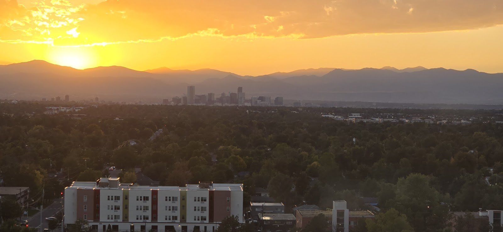 Aerial landscape showing Denver skyline at dusk with snow-covered Rocky Mountains and golden sky