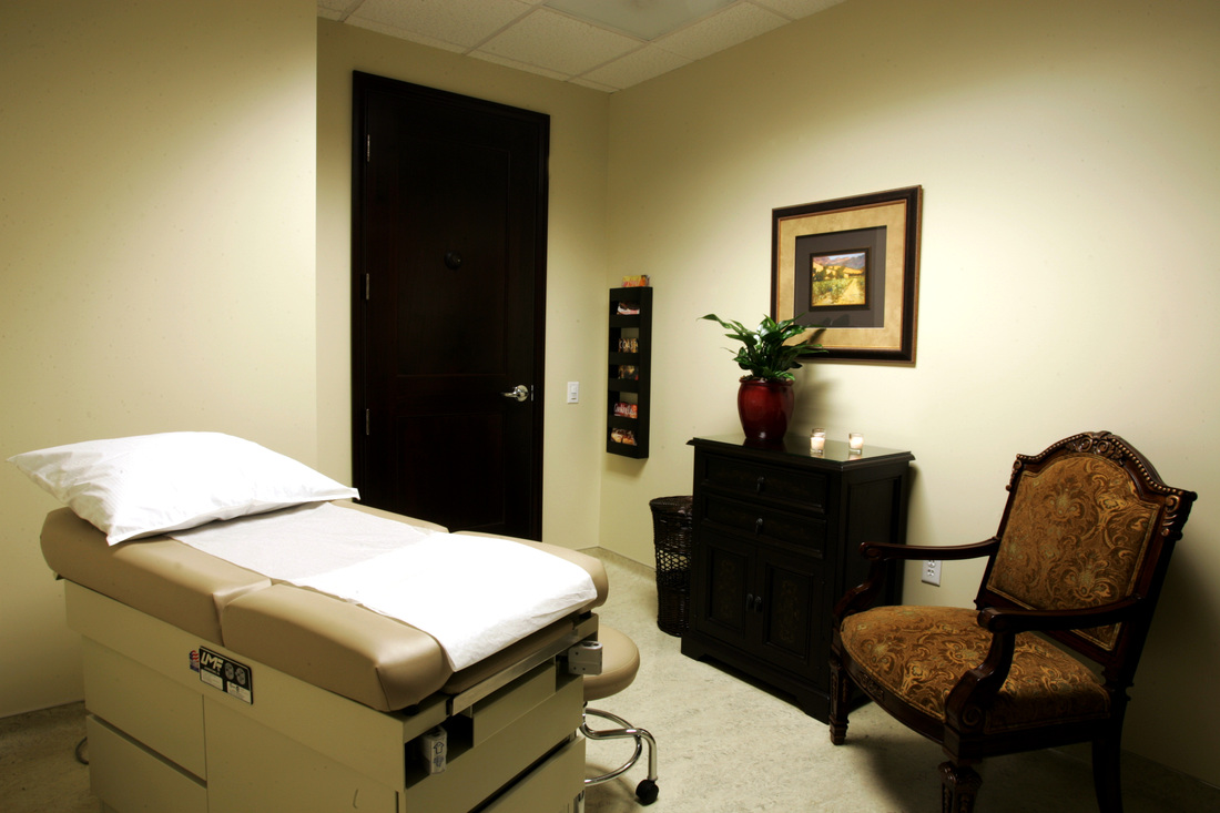 Interior treatment room with massage table, comfortable seating, framed artwork, and pale yellow walls