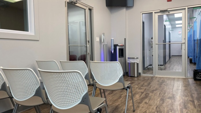 Interior waiting room with white contemporary chairs, glass office partitions, wood flooring, and mounted television screen